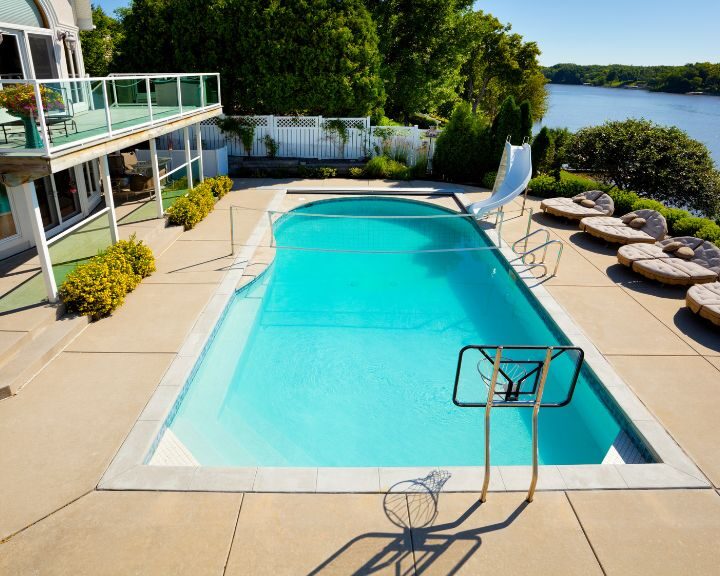 A concrete swimming pool adjacent to a house on a lake in the city.